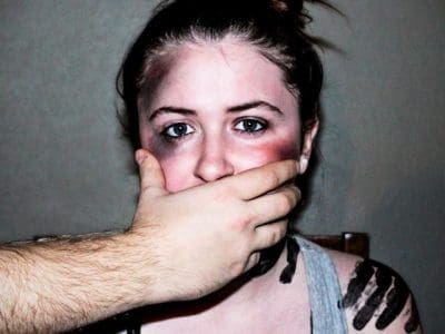 A woman with bruises has a man's hand over her mouth.