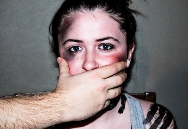 A woman with bruises has a man's hand over her mouth.