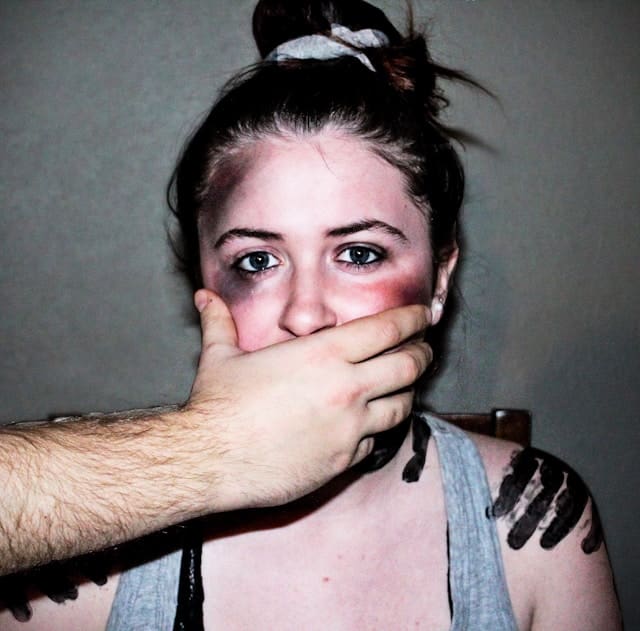 A woman with bruises has a man's hand over her mouth.