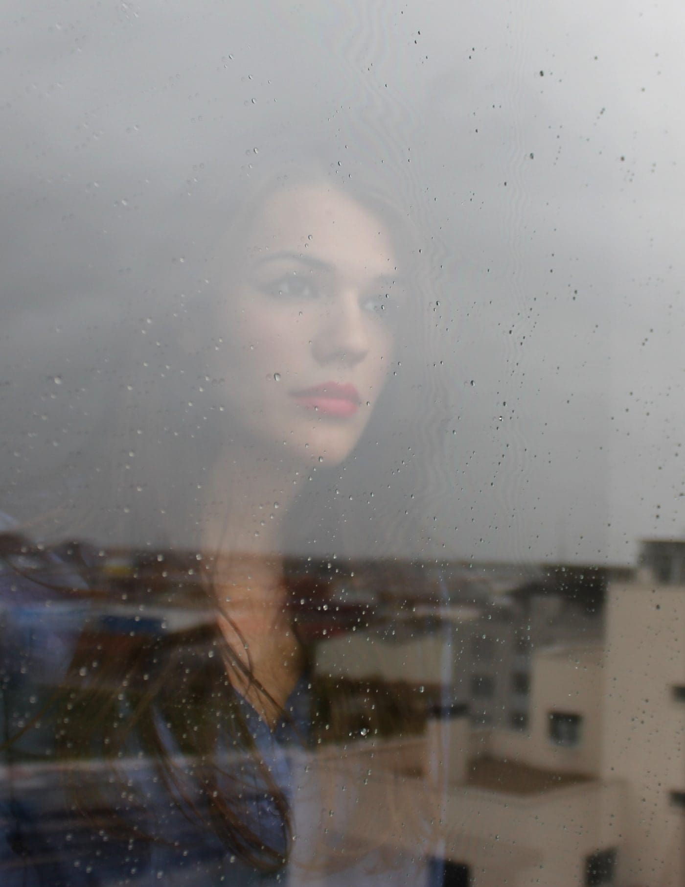 A woman with pink lipstick looks out a rain-streaked window.