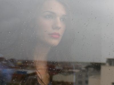 A woman with pink lipstick looks out a rain-streaked window.