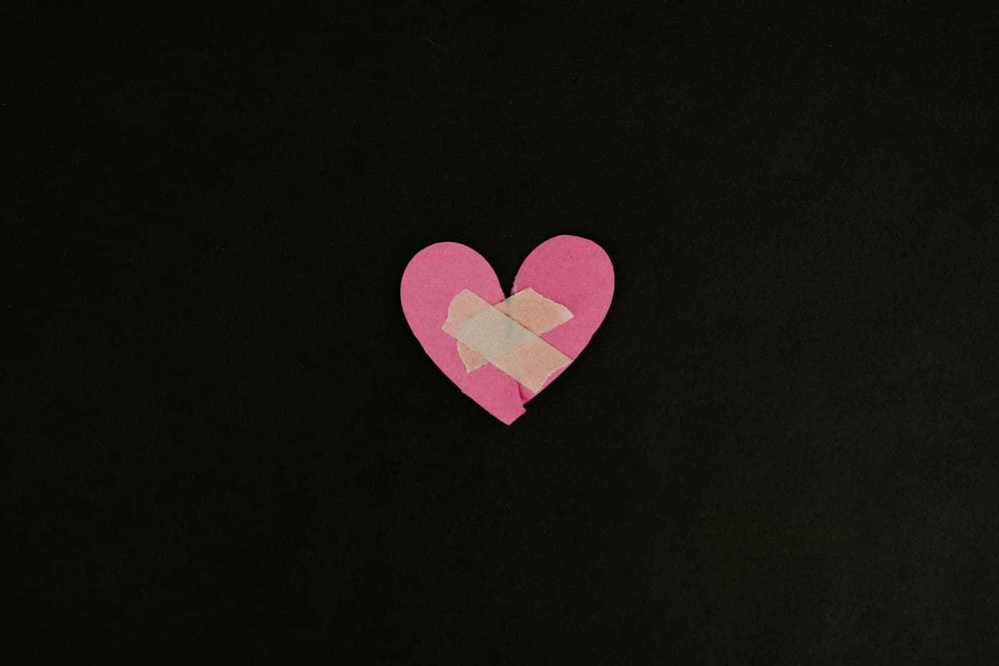 A pink paper heart with a bandage on it against a dark background.