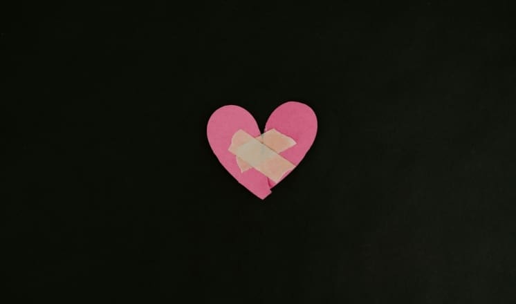 A pink paper heart with a bandage on it against a dark background.
