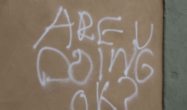 "ARE U DOING OK?" is spray-painted on a tan wall.